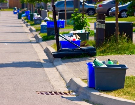 A street scene featuring a series of blue and grey recycling and waste bins placed along the pavement, with some bins open and containing various debris. In the foreground, a grey plastic container holds green and white plastic items, possibly household or garden waste. To the left, a black handheld broom with a long handle rests against the curb, ready for cleaning or collection. The sidewalk is adjacent to a grassy verge with small trees and bushes, and parked cars are visible further along the street. The overall scene suggests an area scheduled for rubbish collection or garden clearance, with belongings arranged for removal services. The environment appears to be in a suburban residential area, likely within the vicinity of Wood Green or nearby in North London, consistent with the local context of Garden Clearance Wood Green's services.