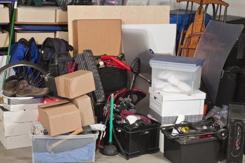 An indoor storage area filled with various household and garden waste items ready for rubbish removal, including cardboard boxes, plastic storage containers, a black tool case, and a vacuum cleaner. Several boxes and bags are stacked on top of each other, some on a white plastic chest and others beside a metal cart or shelves. A red-handled tool or gardening equipment is visible among the clutter. The background features a grey fabric chair, a wooden dining chair, and a shelving unit containing more items. Natural light illuminates the space, highlighting the different textures and colors of the materials, mainly in neutral tones with hints of red and black, creating an organized but cluttered scene typical of a garden clearance or waste collection at a property in the Wood Green area. This image relates to services offered by Garden Clearance Wood Green, specialising in rubbish removal on suitable sites around London and the N22 postcode.
