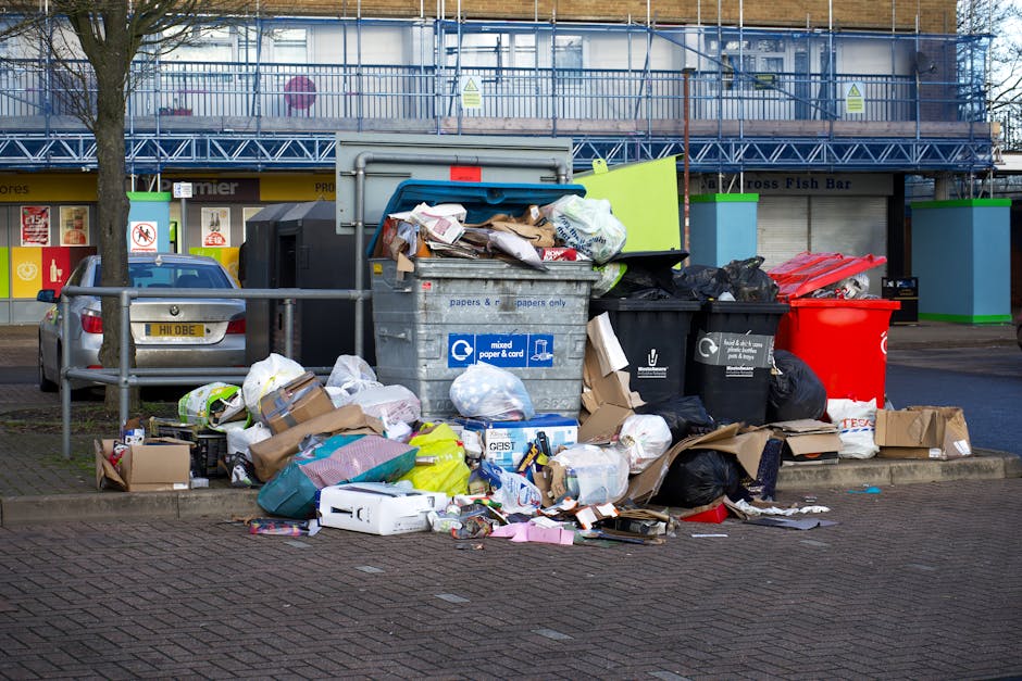 A green wheeled waste container filled with multiple red and black plastic bags containing garden or household waste, situated next to a dark brick wall on a paved surface. The container is positioned
