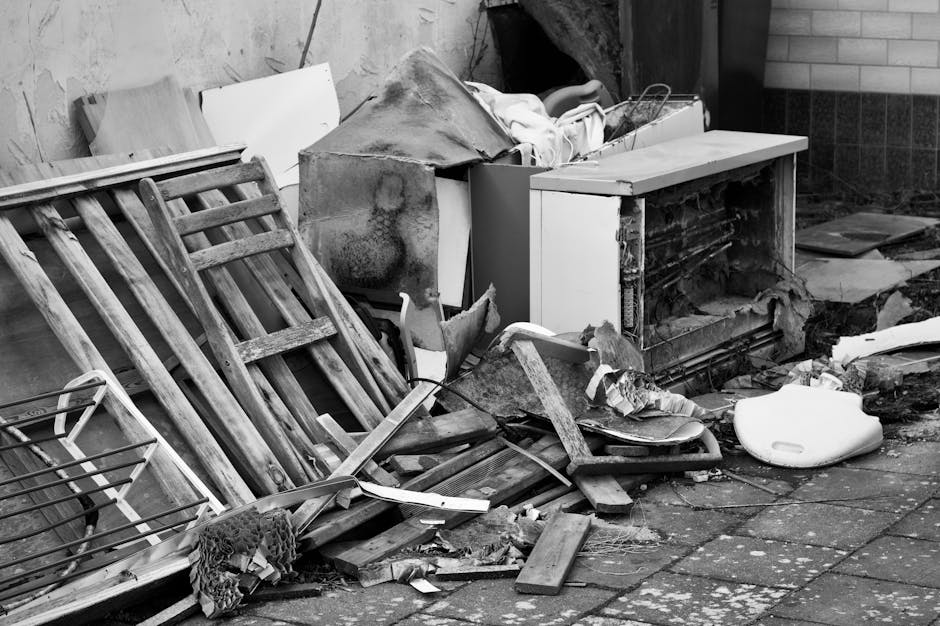 A pile of discarded household waste and debris is shown outside on a paved surface next to a brick wall. The pile includes wooden furniture parts such as slatted bed frames or shelving units, with vis