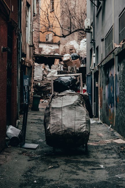 A man with curly dark hair, wearing a black t-shirt and plaid shorts, leans over and inspects a large, rusty, white skip filled with various construction and demolition waste materials. The waste incl